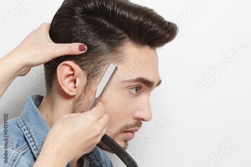 Portrait of handsome young man with black hair having haircut with blade by hairdresser over white background in studio.