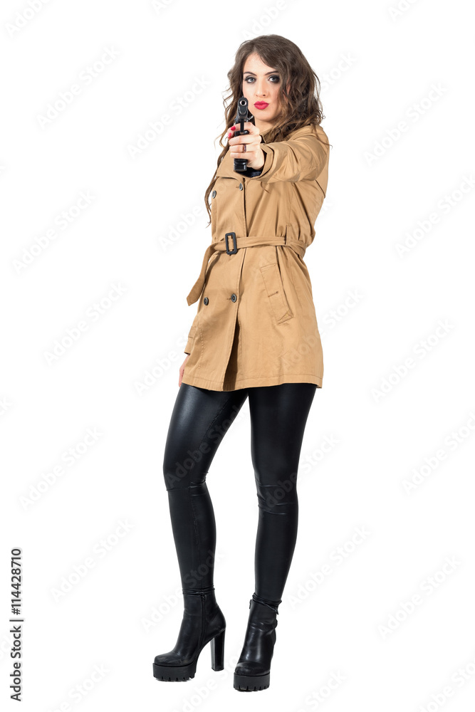 Sexy dangerous wavy hair spy wearing coat aiming weapon at camera. Full body length portrait isolated over white studio background Sexy dangerous wavy hair spy wearing coat aiming weapon at camera. Full body length portrait isolated over white studio background