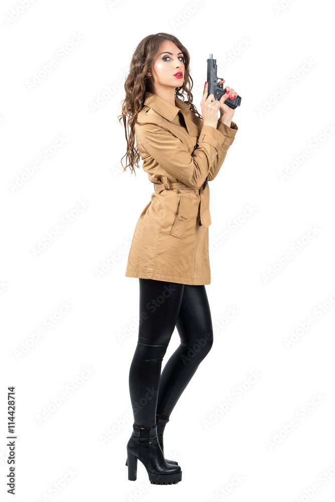 Sexy female spy wearing coat cocking handgun side view. Full body length portrait isolated over white studio background Sexy female spy wearing coat cocking handgun side view. Full body length portrait isolated over white studio background