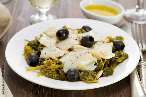cod fish with olives and greens on white plate and glass of wine