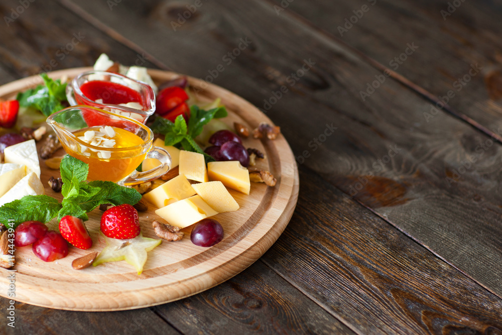 Fruit and cheese plate on wood free space. Top view on round cutting board with different kinds of cheese, fruits and nuts and two sauces. French delicatessen on dark wooden background with copyspace