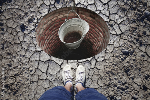 Top view of a man standing in front of built drinking water reservoir with empty metal rusty pail. Conceptual lack of drinking water.