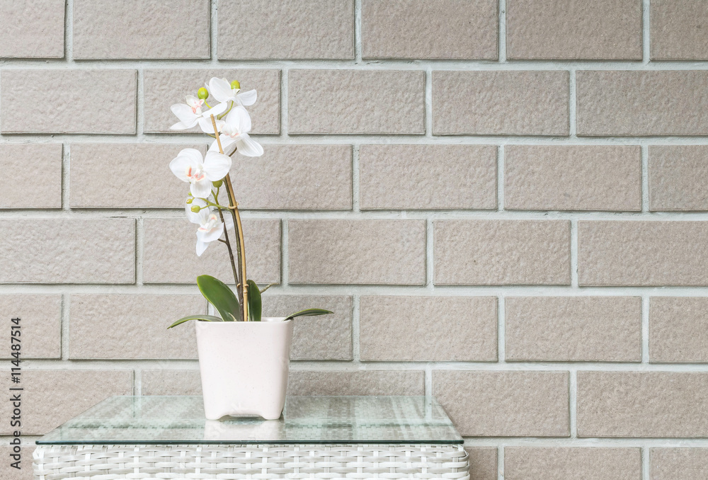 Closeup artificial plant with white orchid flower on pink flower pot on wood weave table on blurred brown brick wall texture background , beautiful interior in relaxation concept of house Closeup artificial plant with white orchid flower on pink flower pot on wood weave table on blurred brown brick wall texture background , beautiful interior in relaxation concept of house