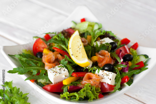 Salmon salad traditional serving with lemon on white wooden background. Top view on white bowl with smoked salmon salad, pepper, brie, olives, lettuce, decorated with slice of lemon.