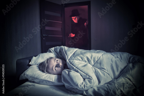 Nightmare. Scared boy lying in the bed while the masked stranger standing in a doorway photo