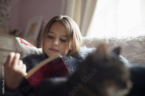 Girl reading book while sitting on sofa with cat