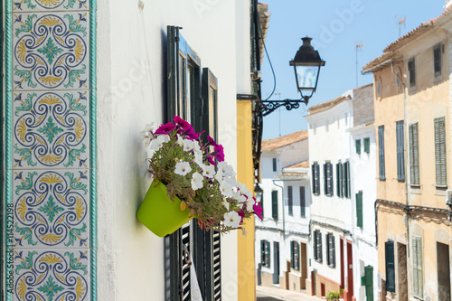 Fototapeta Naklejka Na Ścianę i Meble -  Decorative tiles and flowers in small street at Alaior, Menorca - Close up of tiles with floral pattern and a flowerpot in a small city alley.