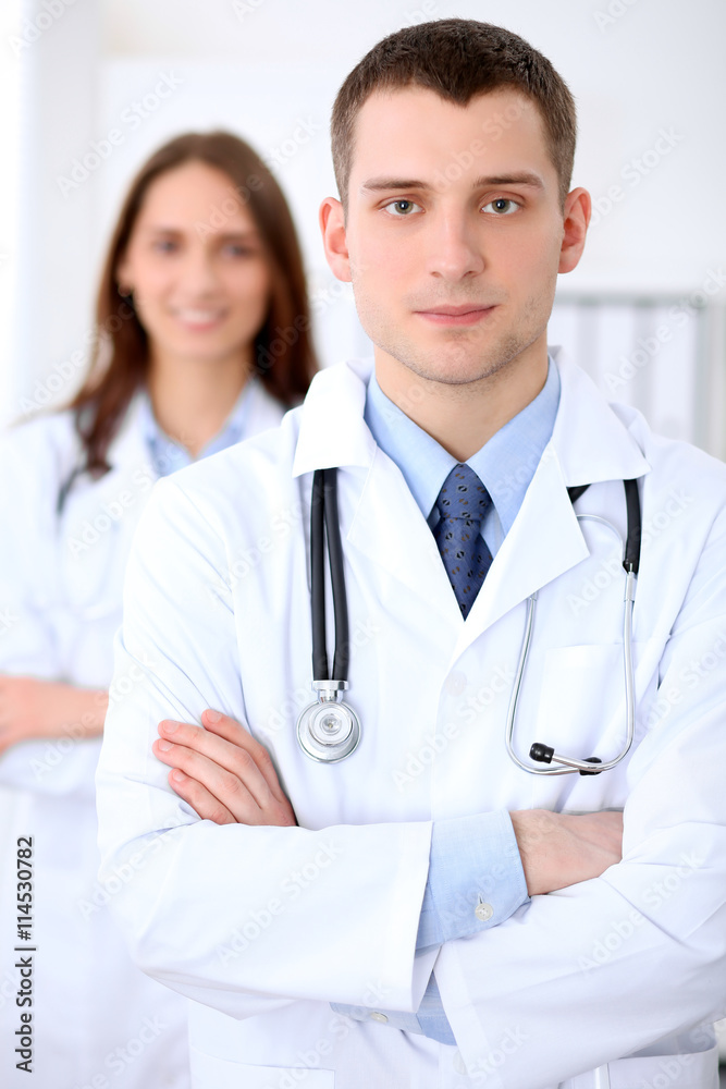 Friendly male doctor on the background of female doctor in hospital office. Ready to examine and help patients. High level and quality medical service concept. Best treatment and patient care concept Friendly male doctor on the background of female doctor in hospital office. Ready to examine and help patients. High level and quality medical service concept. Best treatment and patient care concept