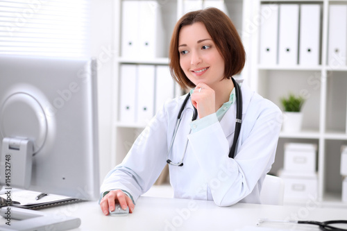 Young brunette female doctor sitting at a desk and working on the computer at the hospital office. Health care, insurance and help concept. Physician ready to examine patient