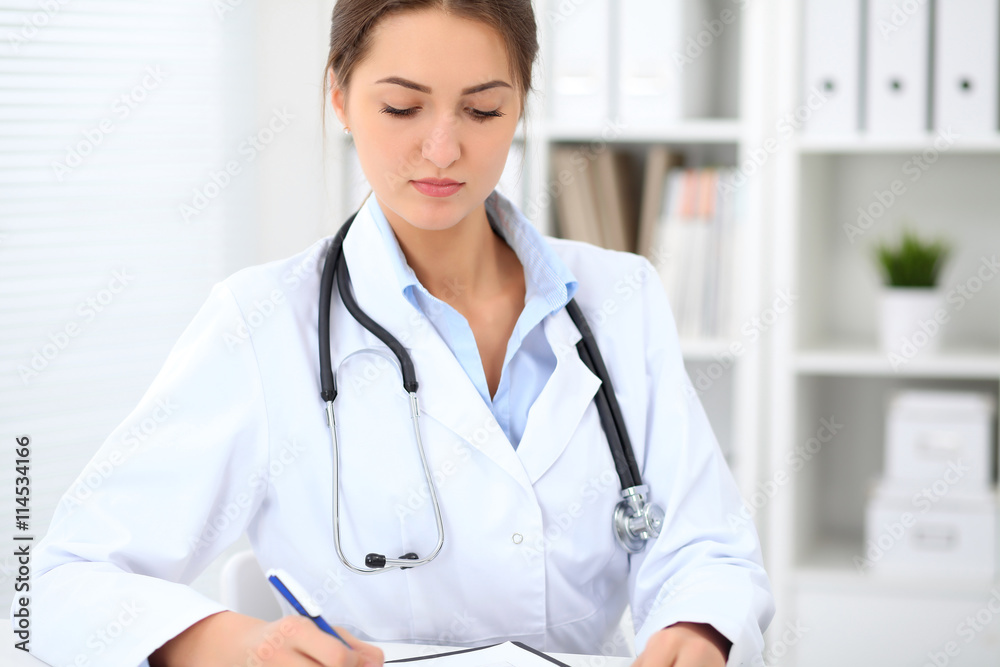 Young brunette female doctor sitting at the table and working at hospital office. Health care, insurance and help concept. Physician ready to examine patient Young brunette female doctor sitting at the table and working at hospital office. Health care, insurance and help concept. Physician ready to examine patient