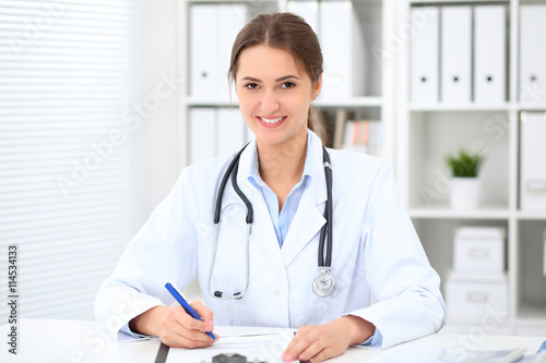 Young brunette female doctor sitting at the table and working at hospital office. Health care, insurance and help concept. Physician ready to examine patient