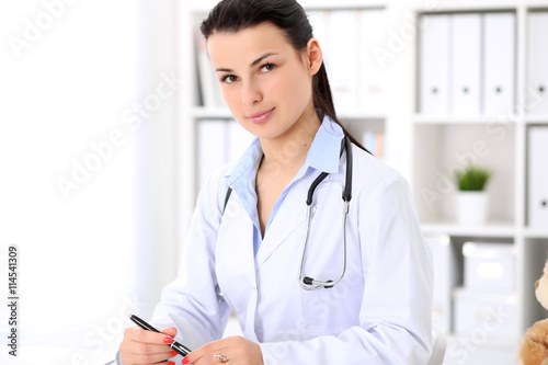 Young brunette female doctor sitting with clipboard near window in hospital and filling up medical history form.