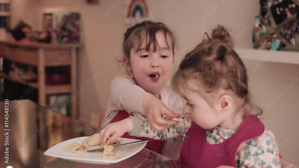 Girls eating pancake from plate at table, mother in background Girls eating pancake from plate at table, mother in background