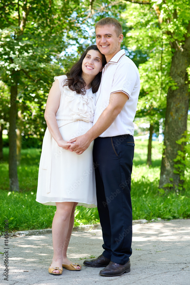 pregnant woman with husband posing in the city park, family portrait, summer season, green grass and trees pregnant woman with husband posing in the city park, family portrait, summer season, green grass and trees