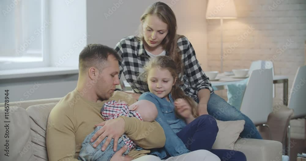 Mother, father and their kids sitting on couch: man holding sleeping baby on his hand, mom and daughter talking to him Mother, father and their kids sitting on couch: man holding sleeping baby on his hand, mom and daughter talking to him