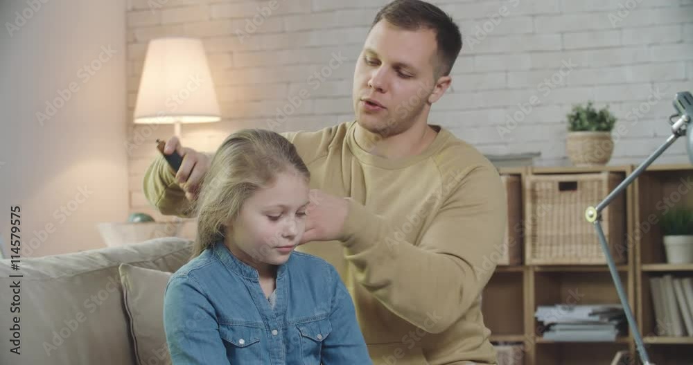 Man brushing hair of his beautiful daughter and talking to her while sitting on sofa in living room Man brushing hair of his beautiful daughter and talking to her while sitting on sofa in living room