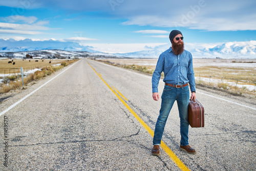 bearded hipster hitch hiker standing in middle of road in nevada with suit case