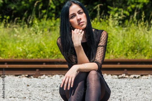 Young beautiful girl in black dress and nylons sitting on rail tracks and daydreaming, green grass and trees in background 