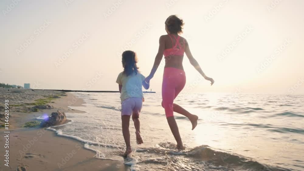 Young mother with her little daugter running on the beach near the water, slowmo