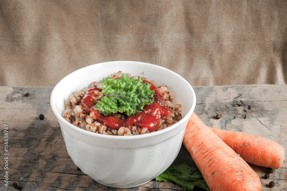 Buckwheat porridge with tomato sauce and leaf salad in a white bowl on a wooden table next to a ceramic bowl with dish is carrots or sliced carrot. Food background. Nice and tasty atmosphere. Buckwheat porridge with tomato sauce and leaf salad in a white bowl on a wooden table next to a ceramic bowl with dish is carrots or sliced carrot. Food background. Nice and tasty atmosphere.
