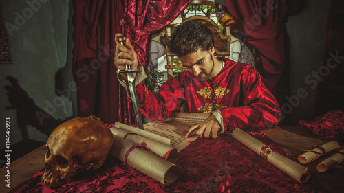 Obraz na plátně Pensive medieval grandee with the dagger in one hand is reading