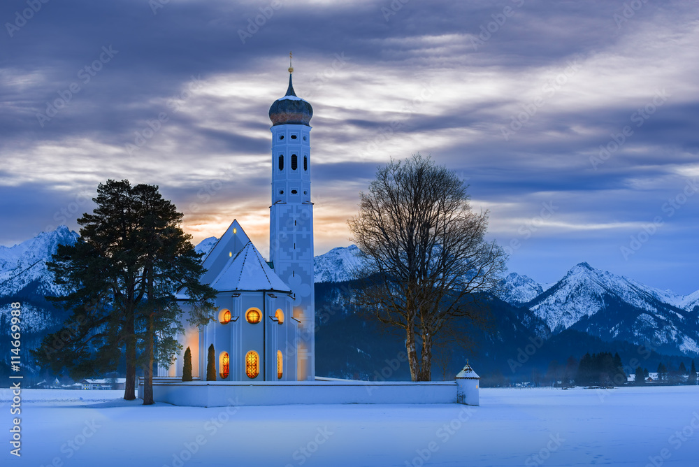 Obraz premium St. Coloman church at dusk on a winter Christmas night with snow and fog and warm candlelight inside