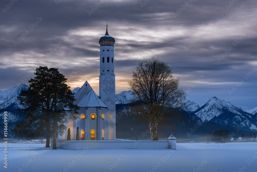 The exterior of the St. Coloman church at dusk on a wintry Christmas evening with warm light coming from the windows