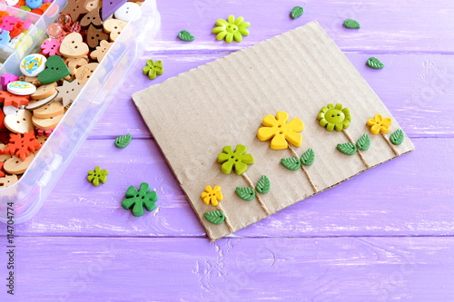 Fun card with flowers. Flowers buttons and leaves on lilac wooden background. Plastic box with colorful buttons. Make kids crafts from recycled materials: cardboard and buttons