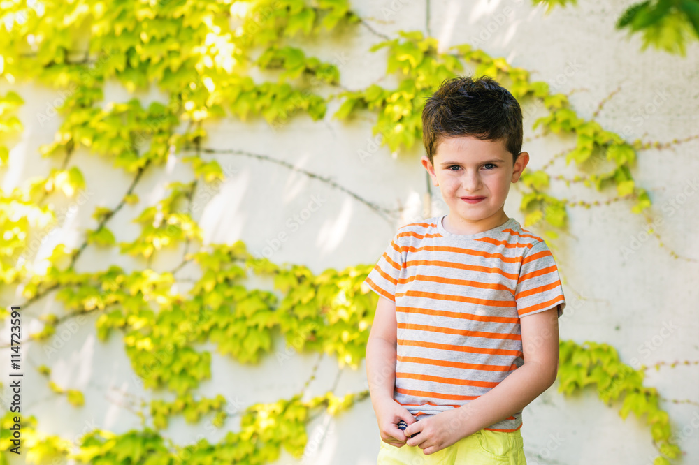 Outdoor portrait of a cute little boy standing against ivy wall on a very sunny day, wearing grey and orange stripe t-shirt Outdoor portrait of a cute little boy standing against ivy wall on a very sunny day, wearing grey and orange stripe t-shirt