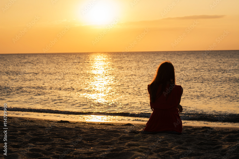 Girl in a red dress at sunrise on a golden beach watching the sea silhouette