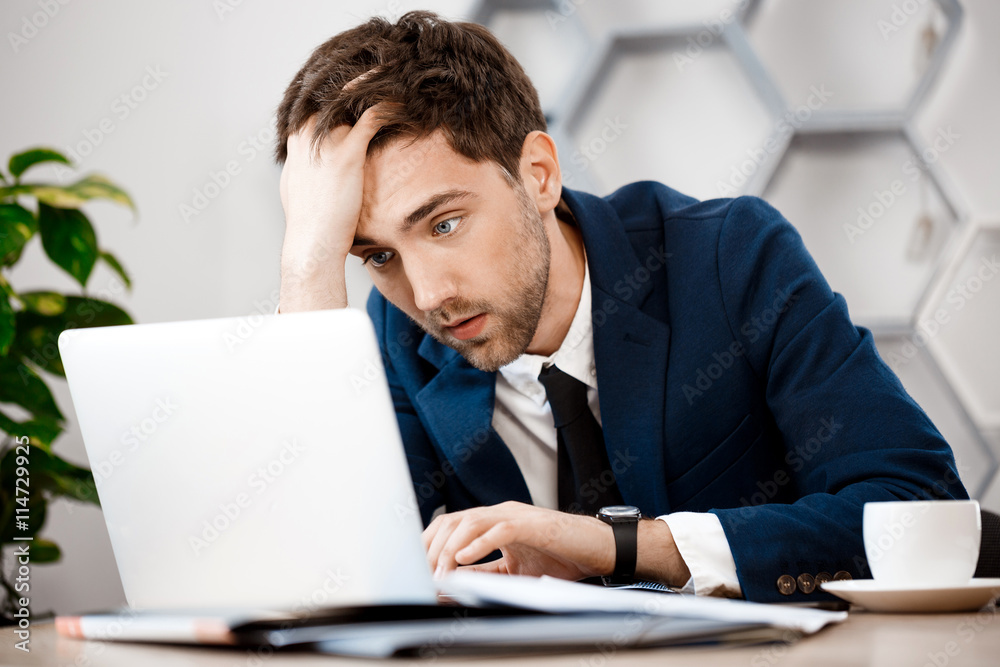 Upset young businessman sitting at laptop, office background. Upset young businessman sitting at laptop, office background.