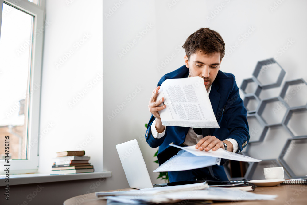 Absentminded young businessman rummaging in papers, office background. Absentminded young businessman rummaging in papers, office background.
