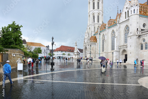 Photography Historic european city square with gothic church and people walking in rain, wet