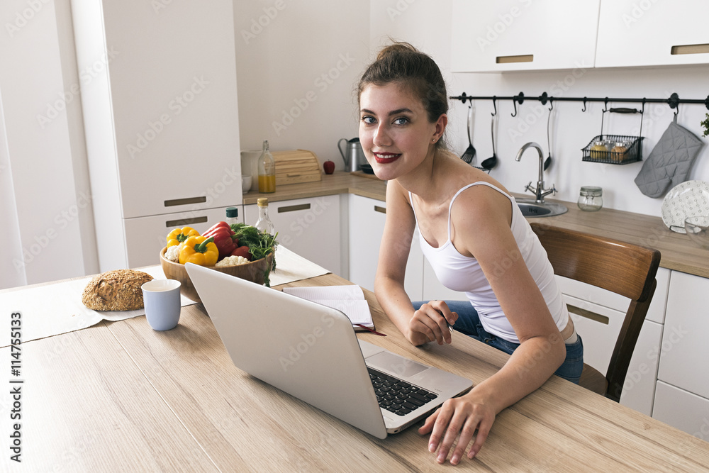 Cheerful housewife smiling at camera at kitchen table with laptop,food and drink Cheerful housewife smiling at camera at kitchen table with laptop,food and drink