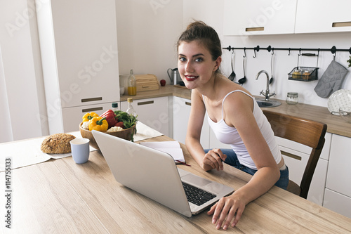 Cheerful housewife smiling at camera at kitchen table with laptop,food and drink