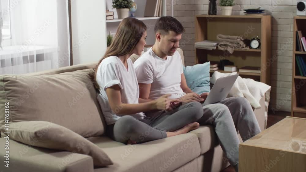 Young boyfriend and girlfriend sitting on the couch and using laptop to make online order with credit card Young boyfriend and girlfriend sitting on the couch and using laptop to make online order with credit card