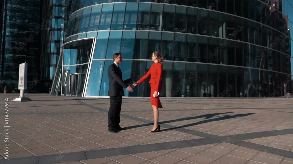 Young cheerful Business partners man and woman doing a handshake in suit outdoor. Glass modern office building district bg. Slow motion wide shot. Teal and orange Young cheerful Business partners man and woman doing a handshake in suit outdoor. Glass modern office building district bg. Slow motion wide shot. Teal and orange