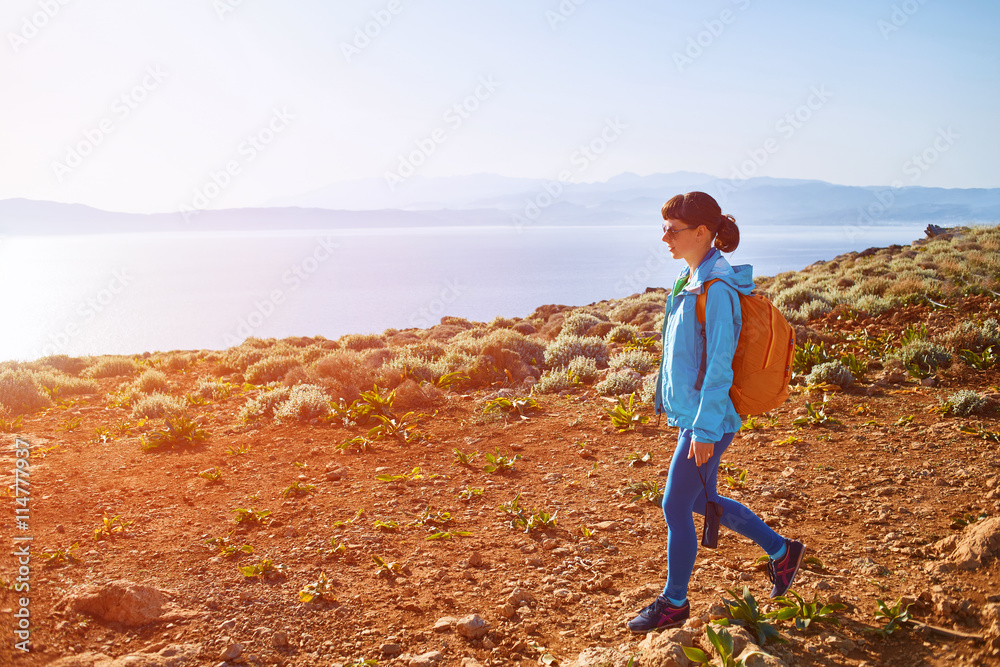 woman traveler with backpack walking on the trail against sea and blue sky at early morning. Balos beach on background, Crete, Greece