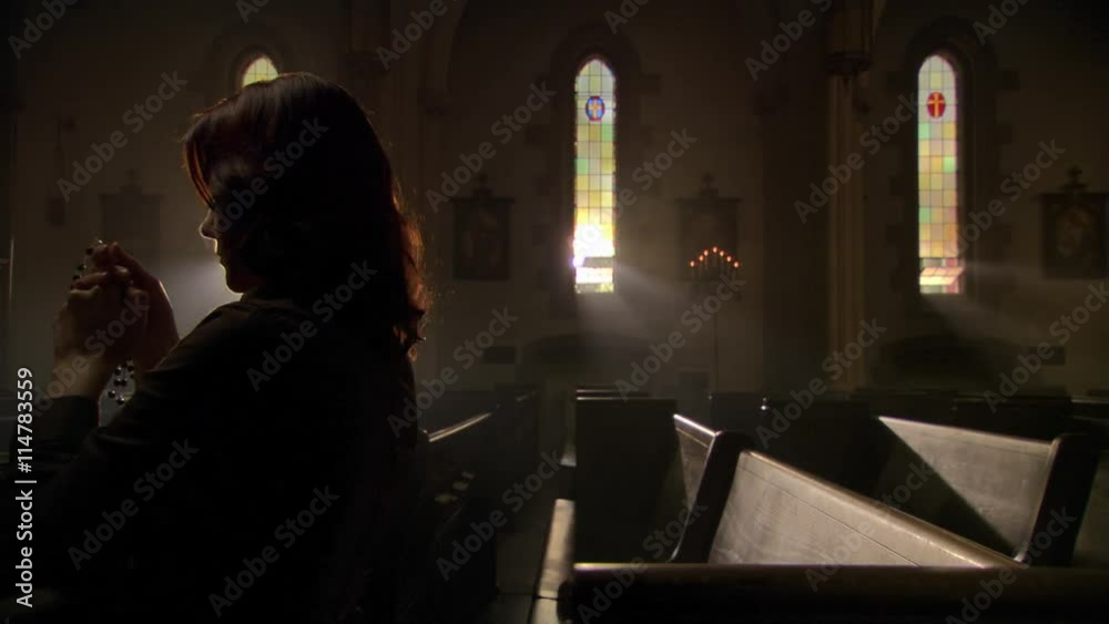Woman in dimly lit church saying prayers with rosary