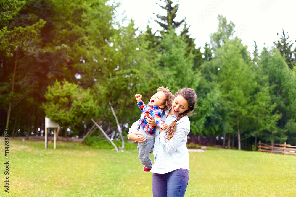 Fototapeta premium Pretty young smiling mother throwing up cheerful child in the park. Woman and toddler son having fun together in summer sunny day. Happy healthy family, love concept,positive human emotions, feelings.