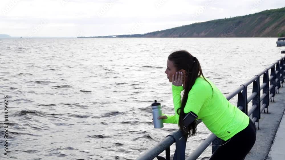 Sport woman runner jogger look by sea. She is exercising and take a break and holding bottle of water on city embankment. View of mountain range is in background. Sport woman runner jogger look by sea. She is exercising and take a break and holding bottle of water on city embankment. View of mountain range is in background.