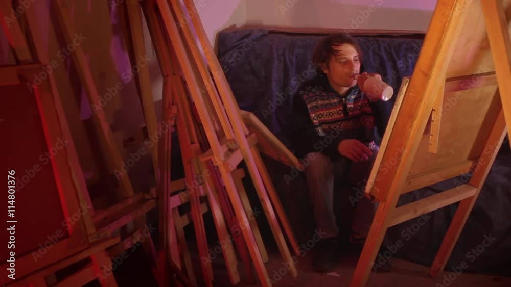 Artist considers his paintings and drinking whiskey in dark workshop wide shot