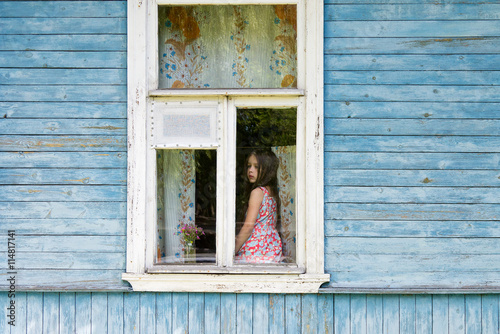Sad bored little girl sitting on the sill and looking out the country house window. Outside view