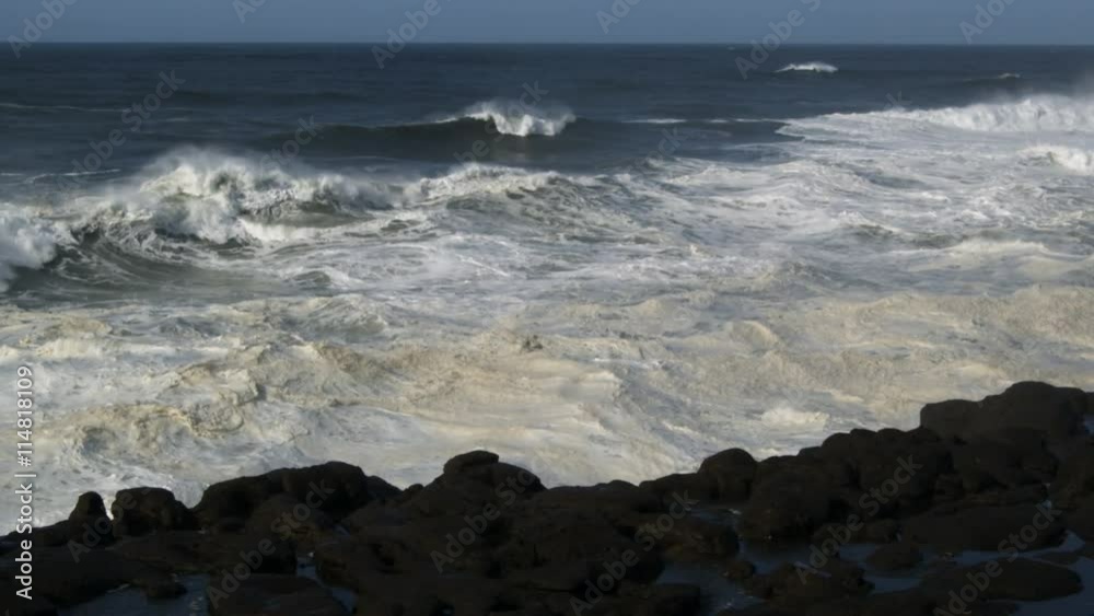 Foaming waves smash over the top of dark rocks along the shore Foaming waves smash over the top of dark rocks along the shore