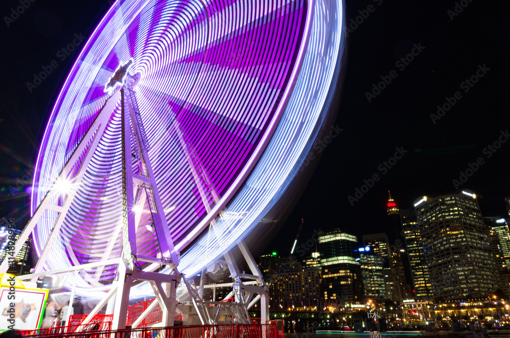Amusement park attractions. Spinning ferris wheel at night with cityscape on the background. Motion blur