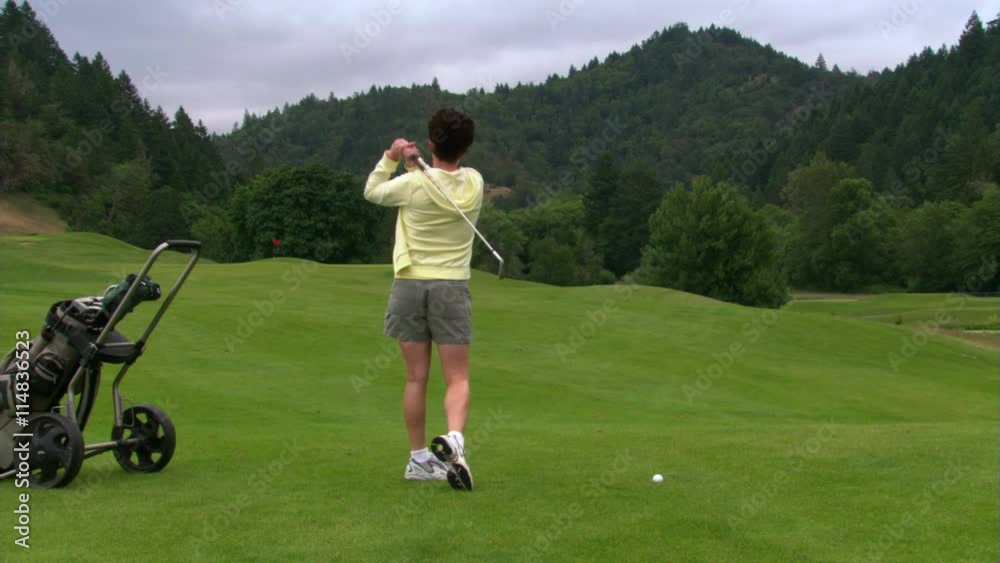 Woman golfer hitting ball, returning club to bag, and walking toward distant flag with bag in tow Woman golfer hitting ball, returning club to bag, and walking toward distant flag with bag in tow