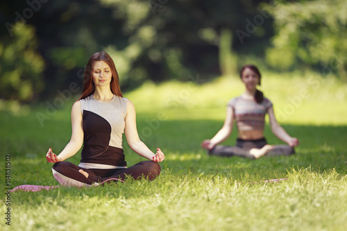 Two girls doing yoga exercises in park. Two young women in sport clothes standing on a green grass and meditate. 
