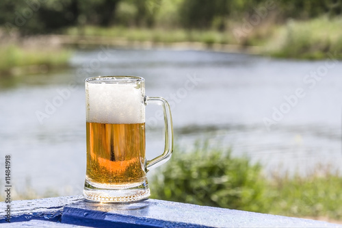 Canvas Print Mug of frothy beer on the table near lake