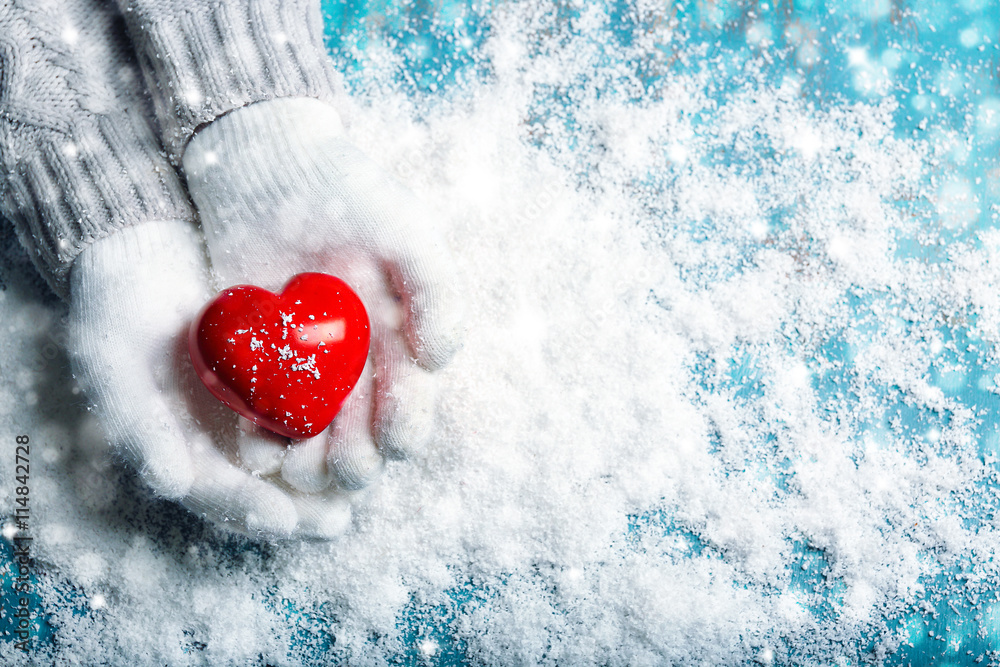 Hands in warm white gloves holding red heart on snowy background. Snow effect