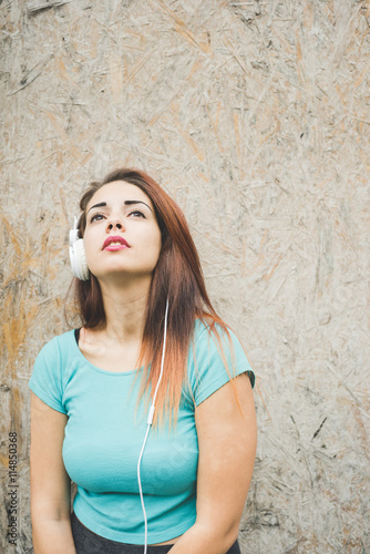 Half length of young handsome caucasian reddish straight hair woman leaning against a wall listening music with headphones, looking upward, pensive - thinking future, thoughtful concept © Eugenio Marongiu